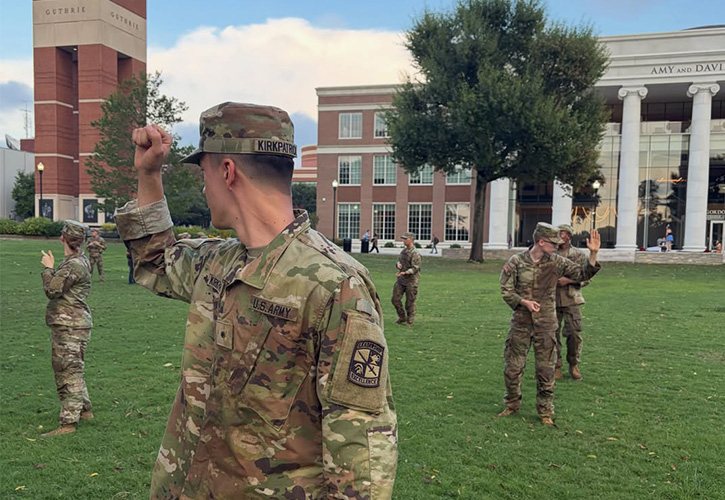 An Army ROTC cadet in the foreground raises a clenched fist during outdoor training on a grassy campus lawn, with fellow cadets practicing hand-and-arm signals across the field. Guthrie Bell Tower and campus buildings stand in the background.