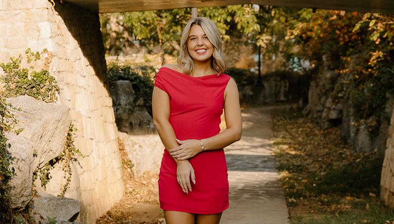 Mikayla Gambrel, a WKU education major, stands smiling brightly under a stone overpass on a sunny day. She is wearing a bright red dress with an asymmetrical neckline, with a paved walkway and green and yellow trees in the background.
