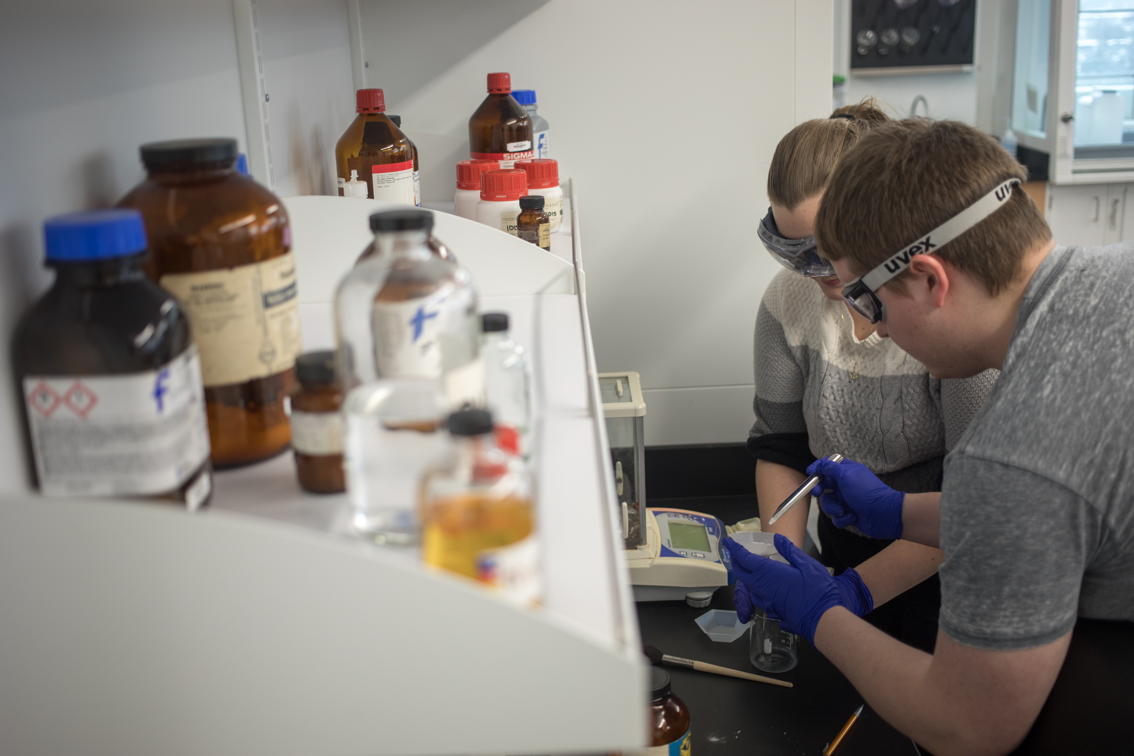 Two students wearing lab goggles weigh materials in a Chemistry lab.