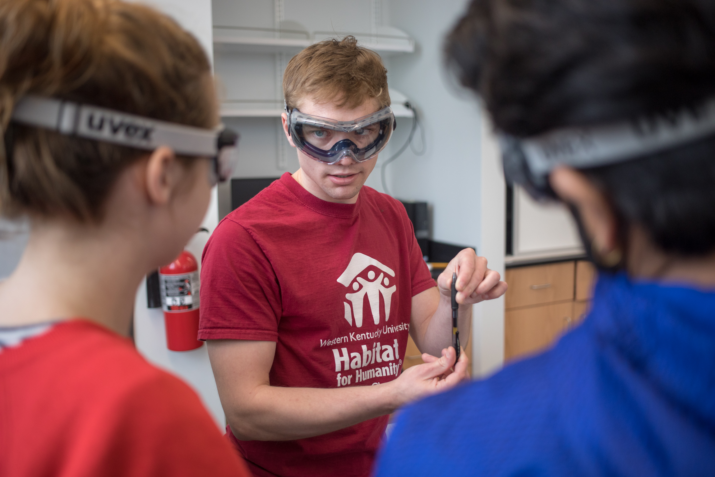 A student wearing lab goggles talks to two other students.