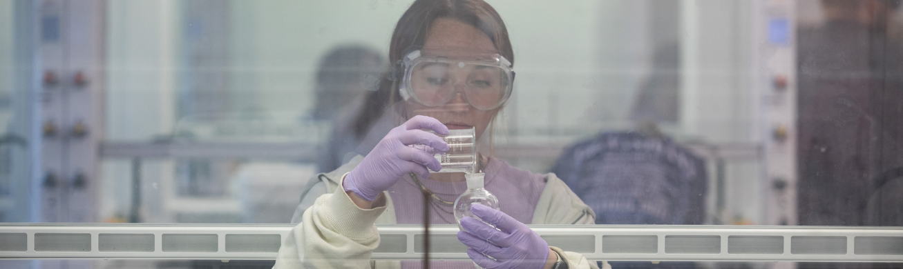 A student wearing goggles pours liquid from a beaker into a separatory funnel underneath a fume hood.