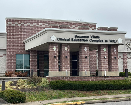 Front entrance of the Suzanne Vitale Clinical Education Complex at WKU. The modern building features red and light-gray patterned brickwork, large glass windows, and a covered drop-off area supported by four heavy brick pillars with star icons framing the building's signage.