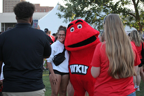 Individual standing next to Big Red, WKU's mascot, while another individual stands nearby and a third individual is taking a photo